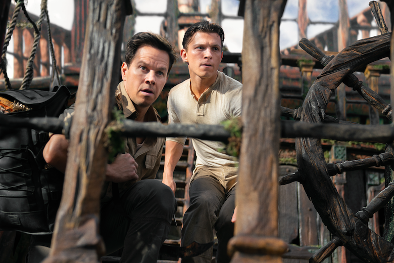 Mark Wahlberg and Tom Holland crouch on a wooden deck, a large wheel nearby.   