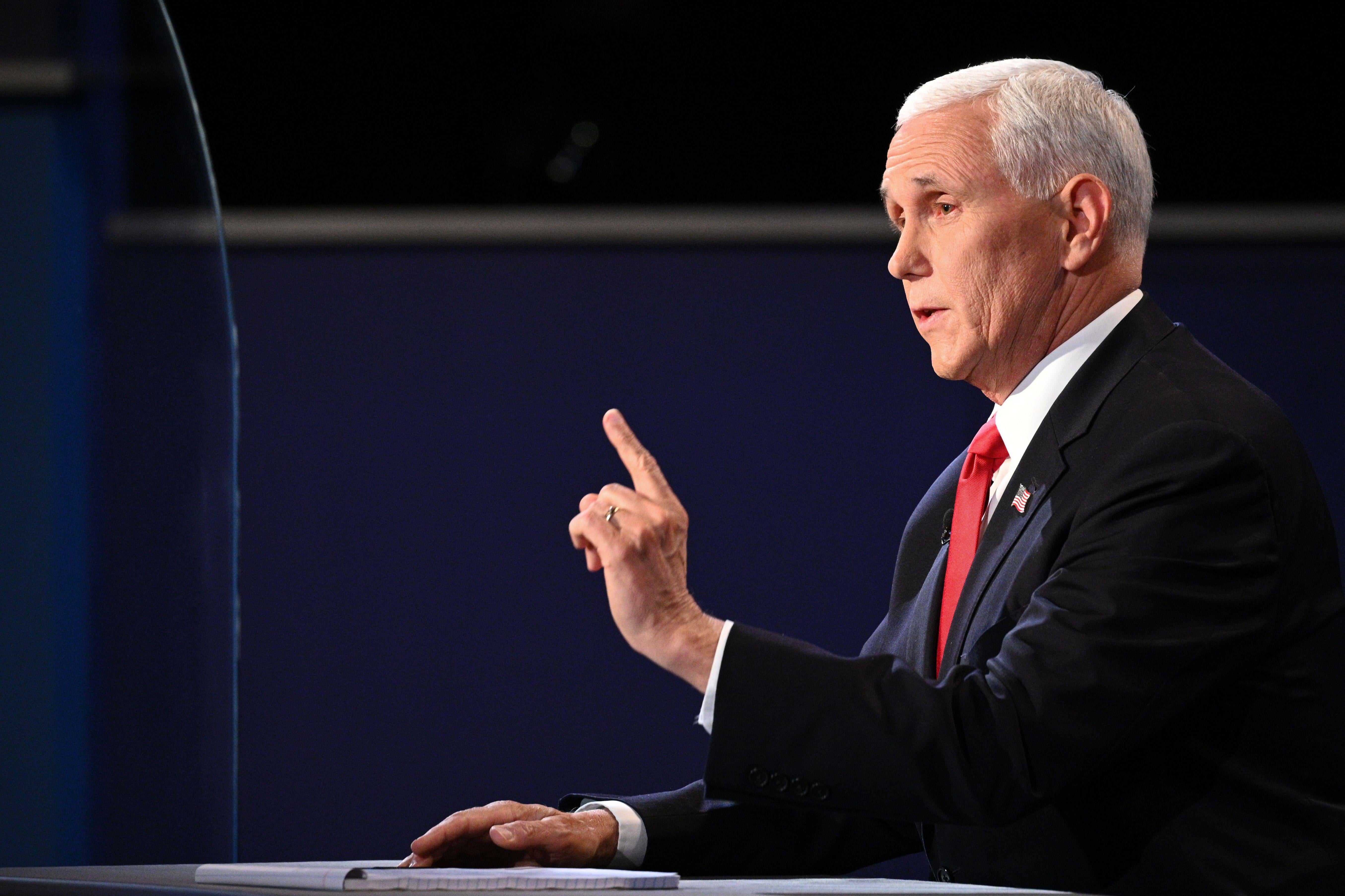Mike Pence, in profile at the right side of the picture, speaks while sitting with his right hand flat on a desk and his left index finger wagging.  