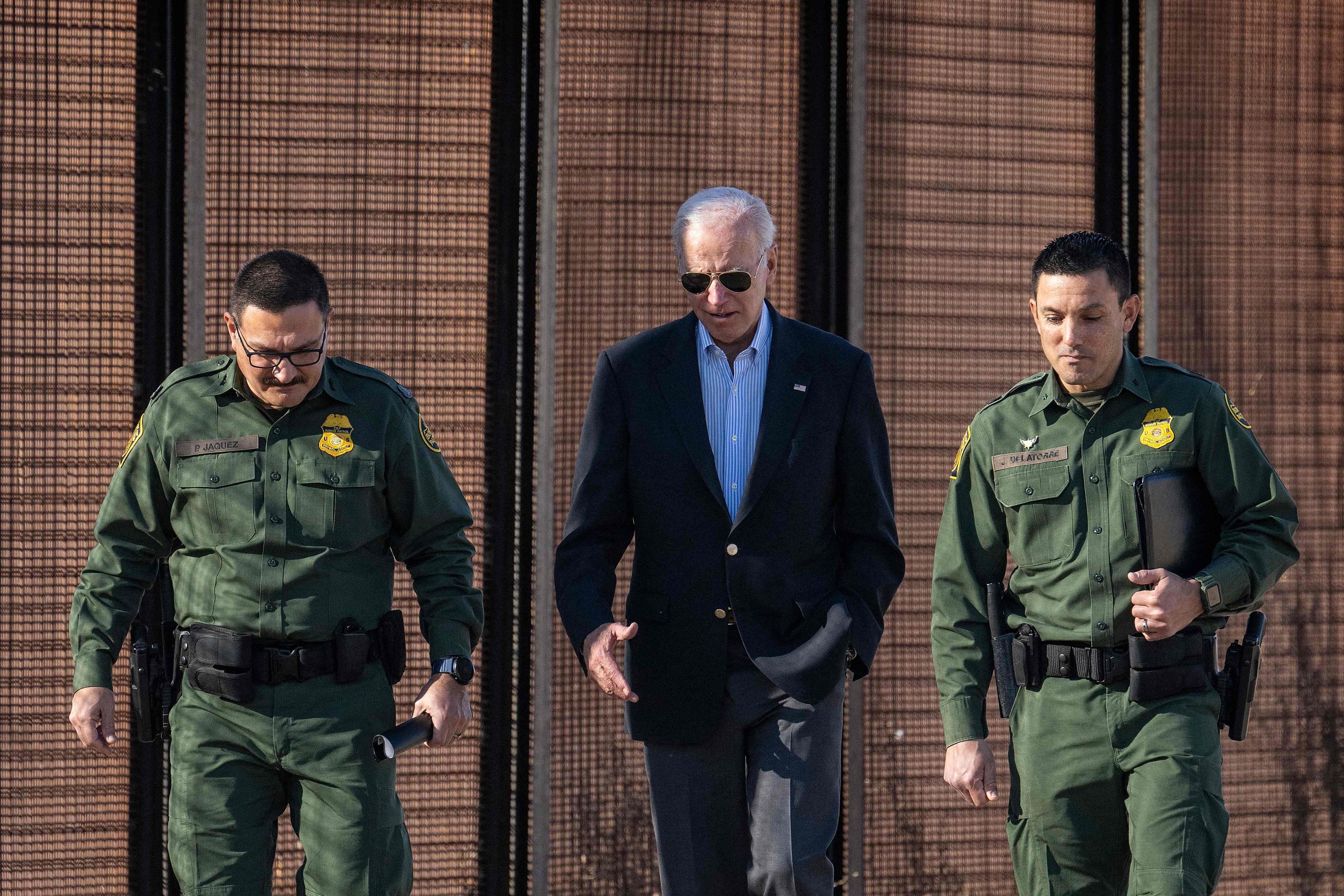 Biden in aviators walking between two uniformed officers with the border fence looming behind them