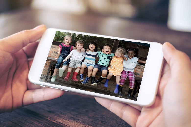 Hands hold an iPhone screen that displays a group of six children sitting together on a bench.