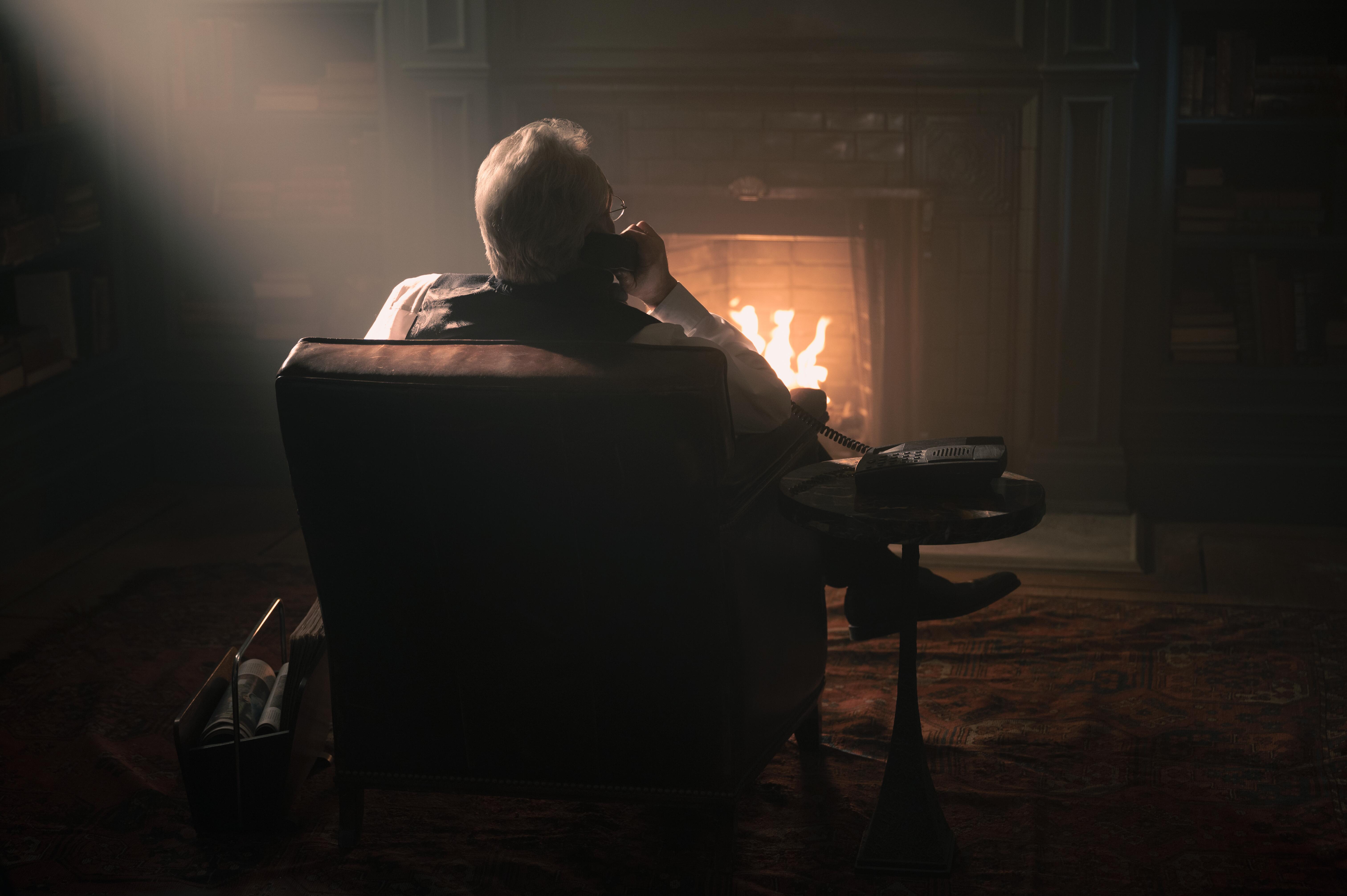 Viewed from behind, a man sits in a leather chair in front of a fire as he talks on a corded phone.