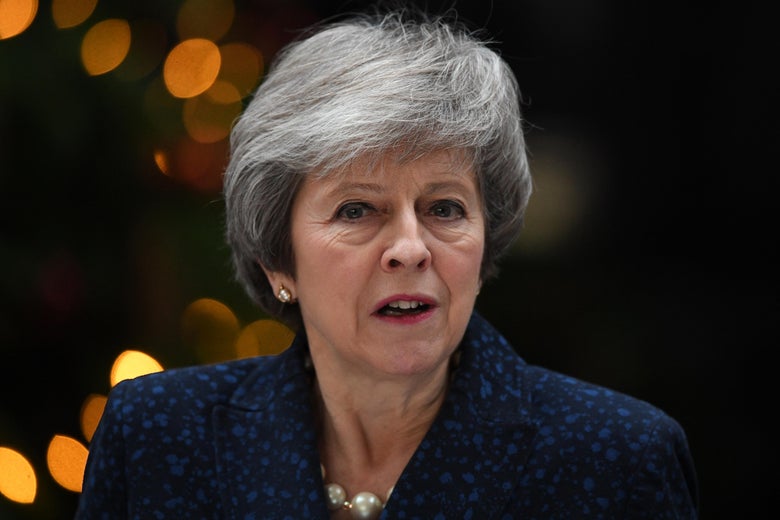 Theresa May speaks and looks toward the camera. Behind her, there appear to be Christmas lights.