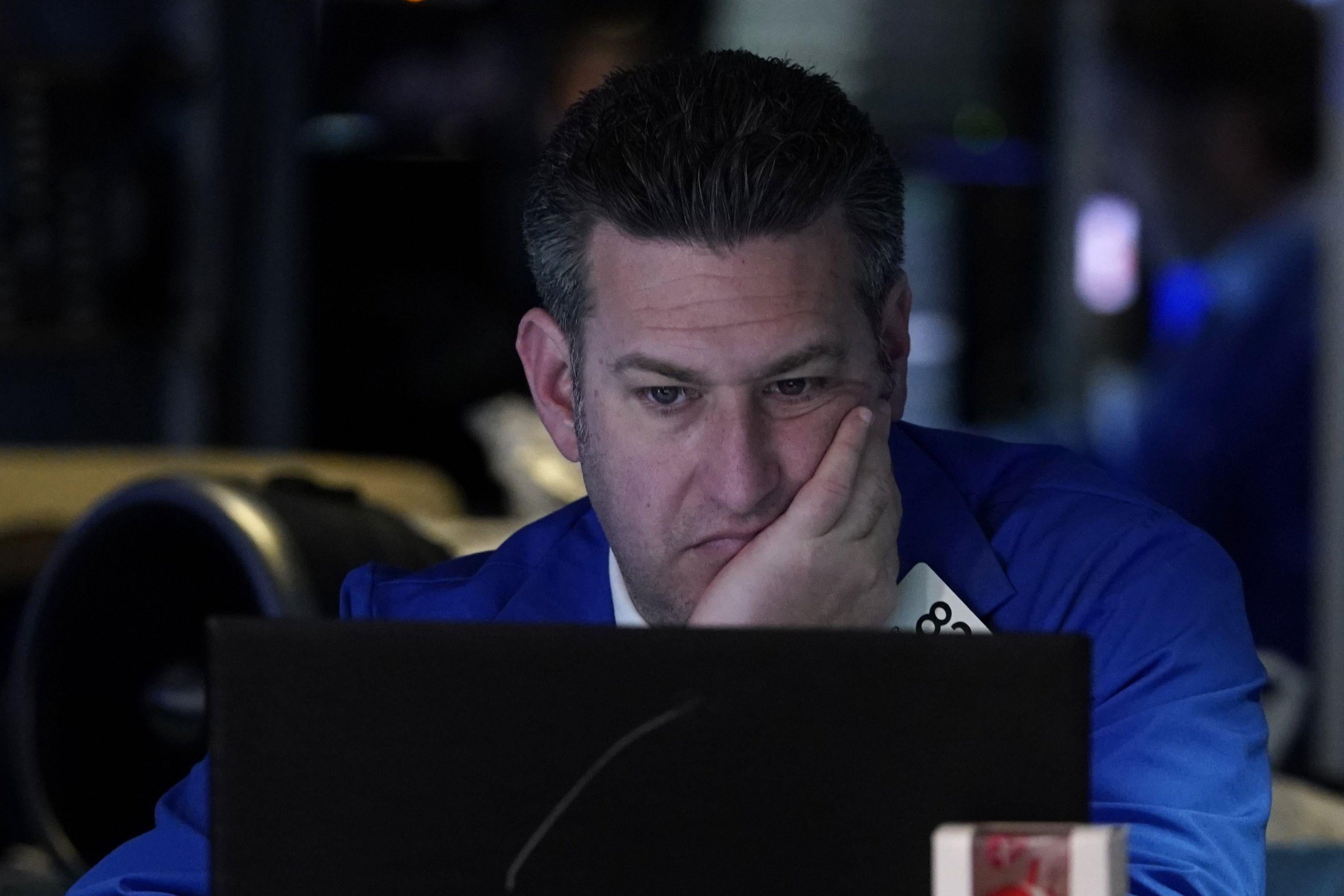 A man in a blue coat sits at a computer with his head in his hand.