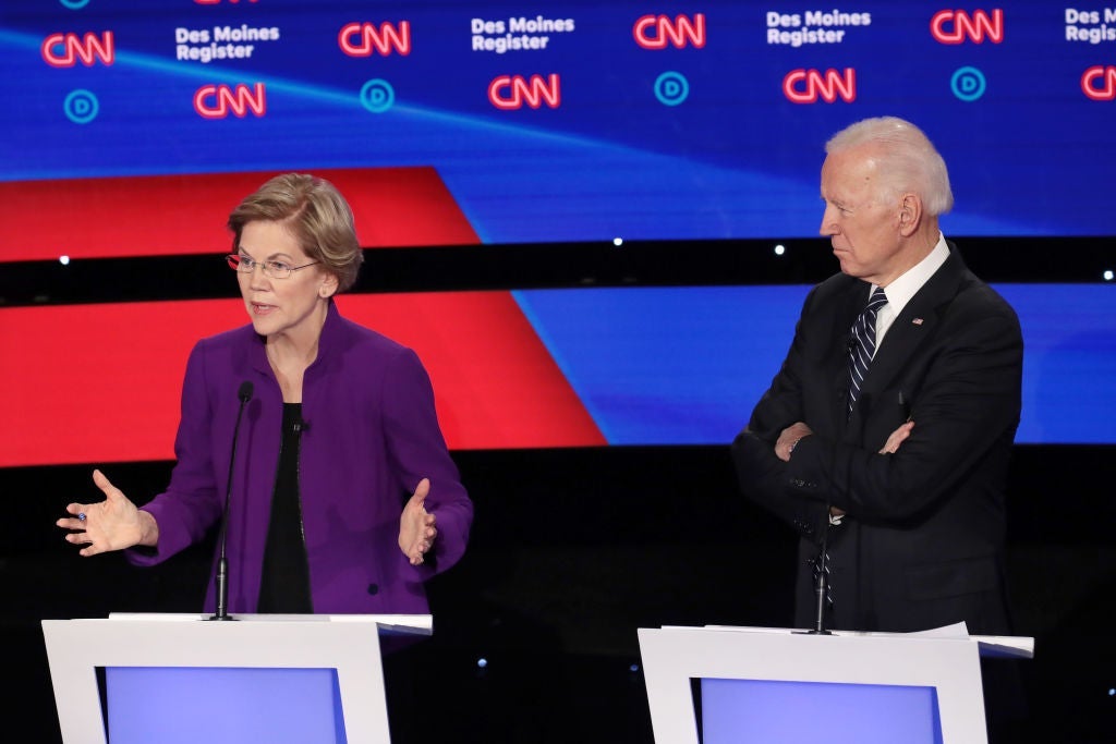 Warren gestures while speaking from her lectern as Biden watches with his arms folded, standing behind the lectern next to hers.