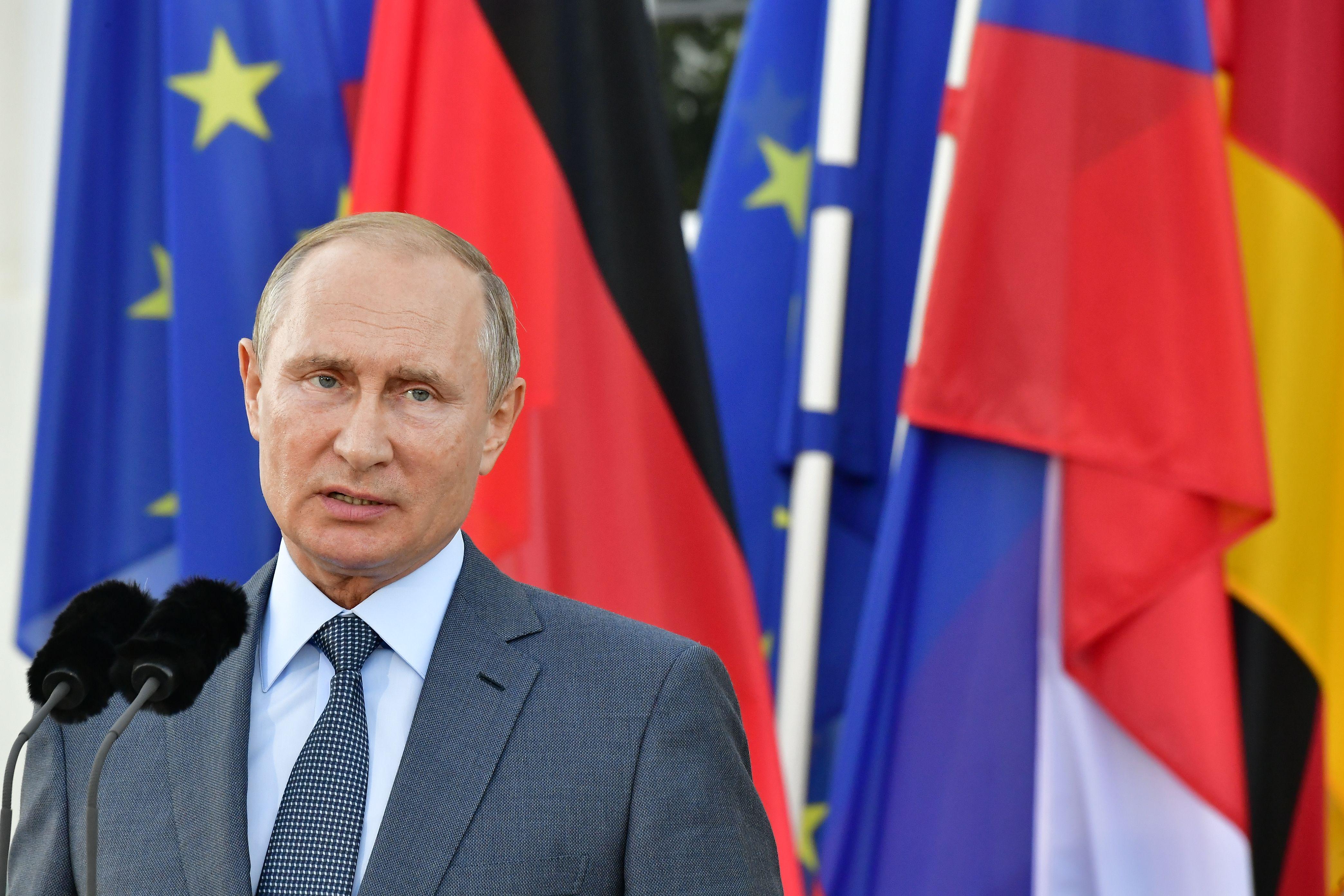 Russian President Vladimir Putin stands behind a microphone and in front of a backdrop of flags