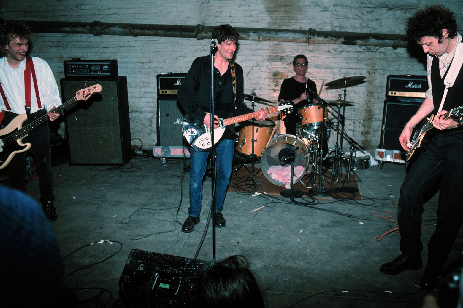 The band members smile while playing on a basement stage against a white brick wall. 