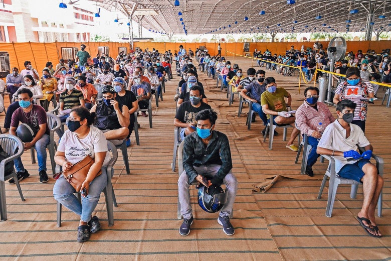 People in masks sit in spaced-out plastic chairs in a big room.