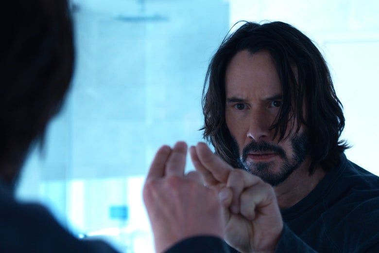 A bearded Neo (Keanu Reeves) taps on a mirror, where he sees himself, looking confused.