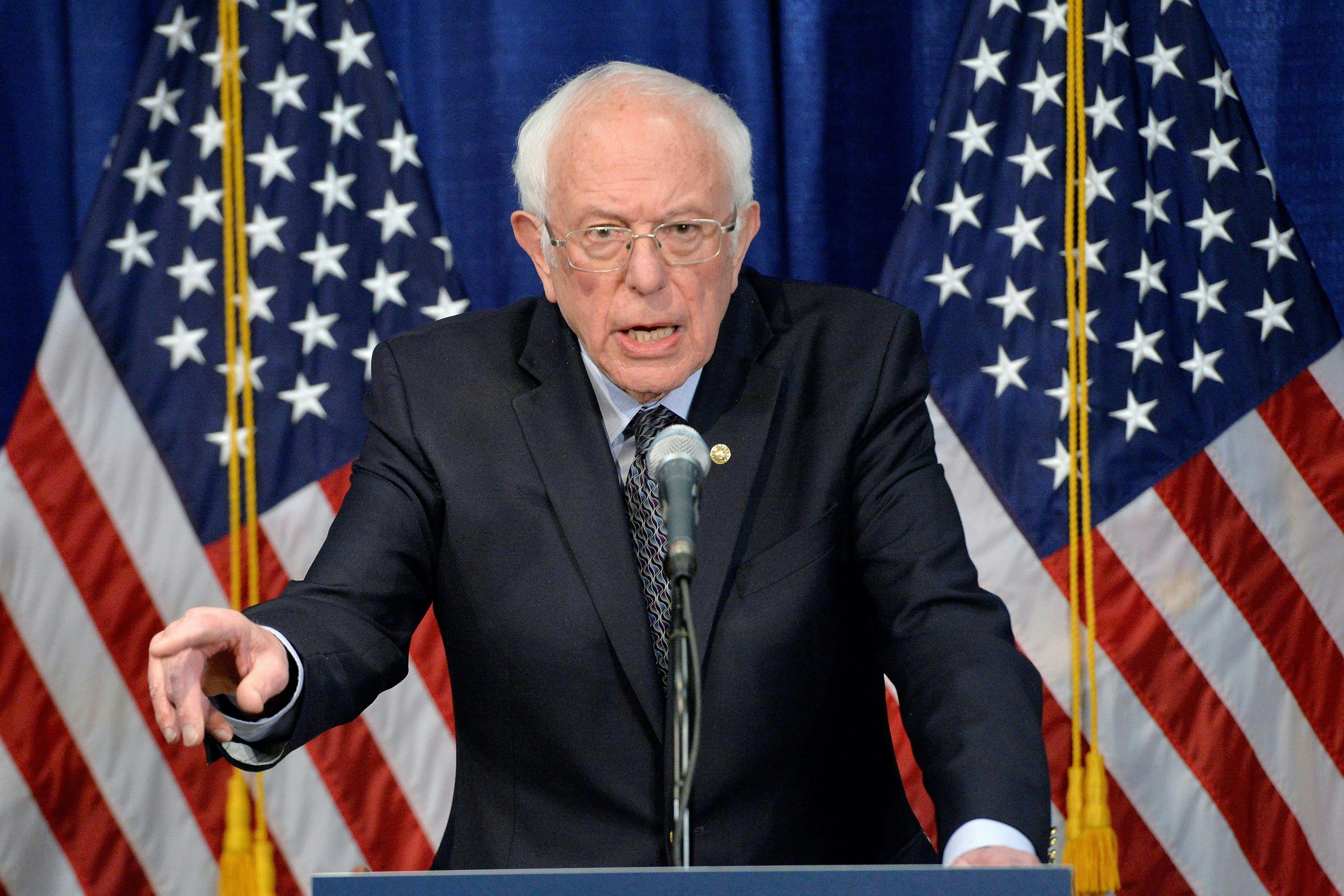 Bernie Sanders speaks at a microphone, in front of two American flags. 