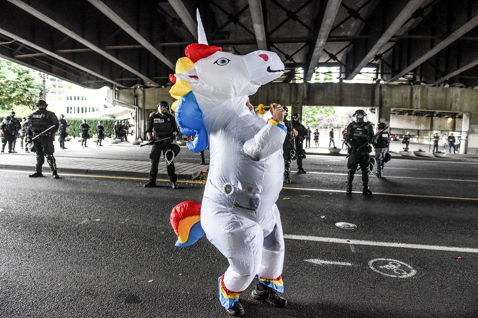 A person in an inflatable unicorn costume stands in a street under an overpass with police in riot gear in the background.