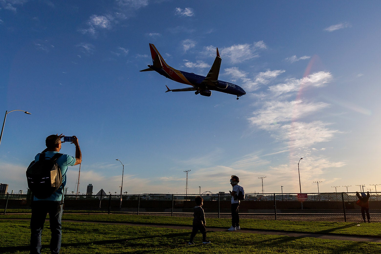 As a Southwest plane descends overhead to land, three individuals stand in a field to watch. One man to the left holds up a smartphone to take a photo of the plane. 