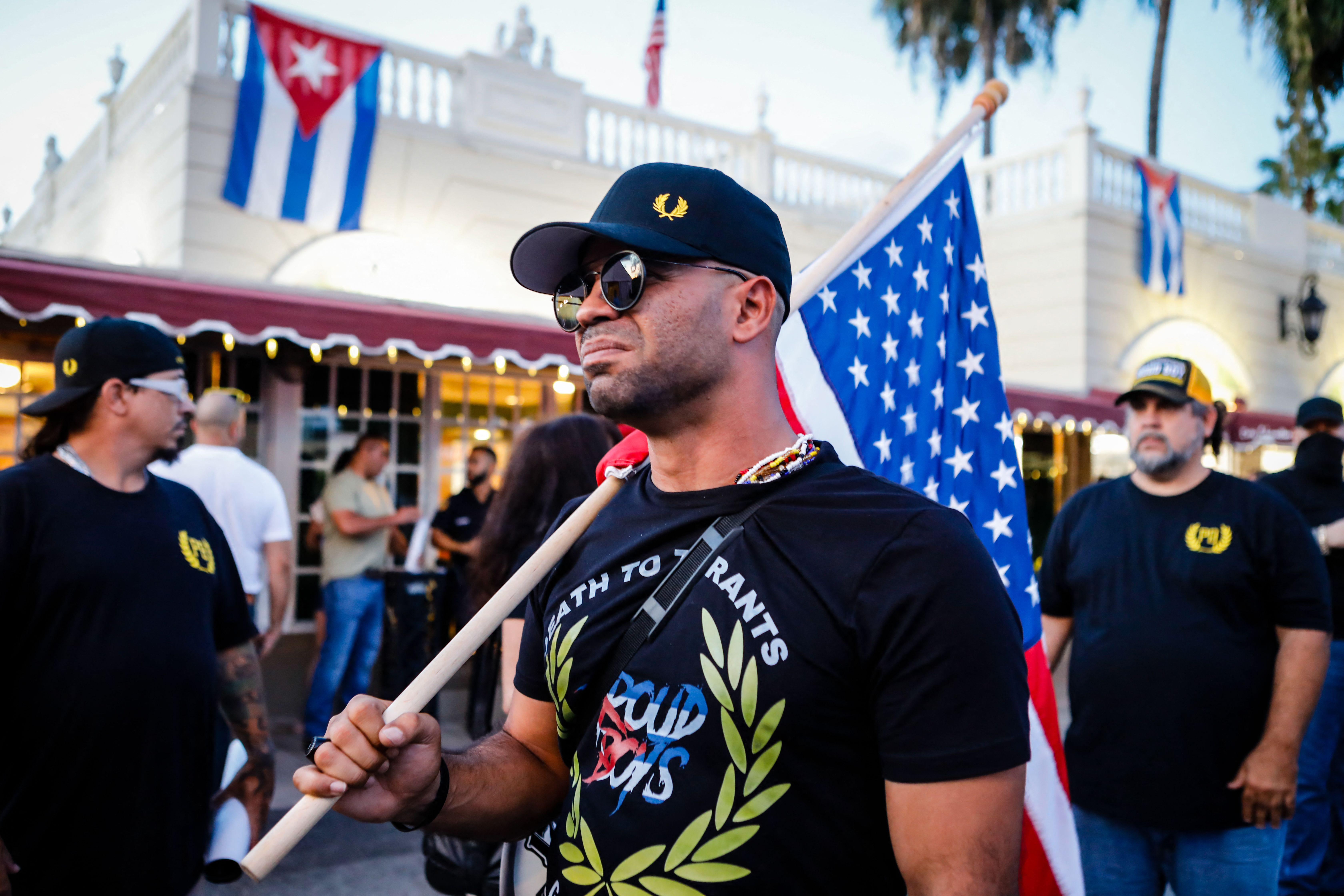 Tarrio wearing a Proud Boys T-shirt and holding an American flag over his shoulder as he marches in a street during a protest