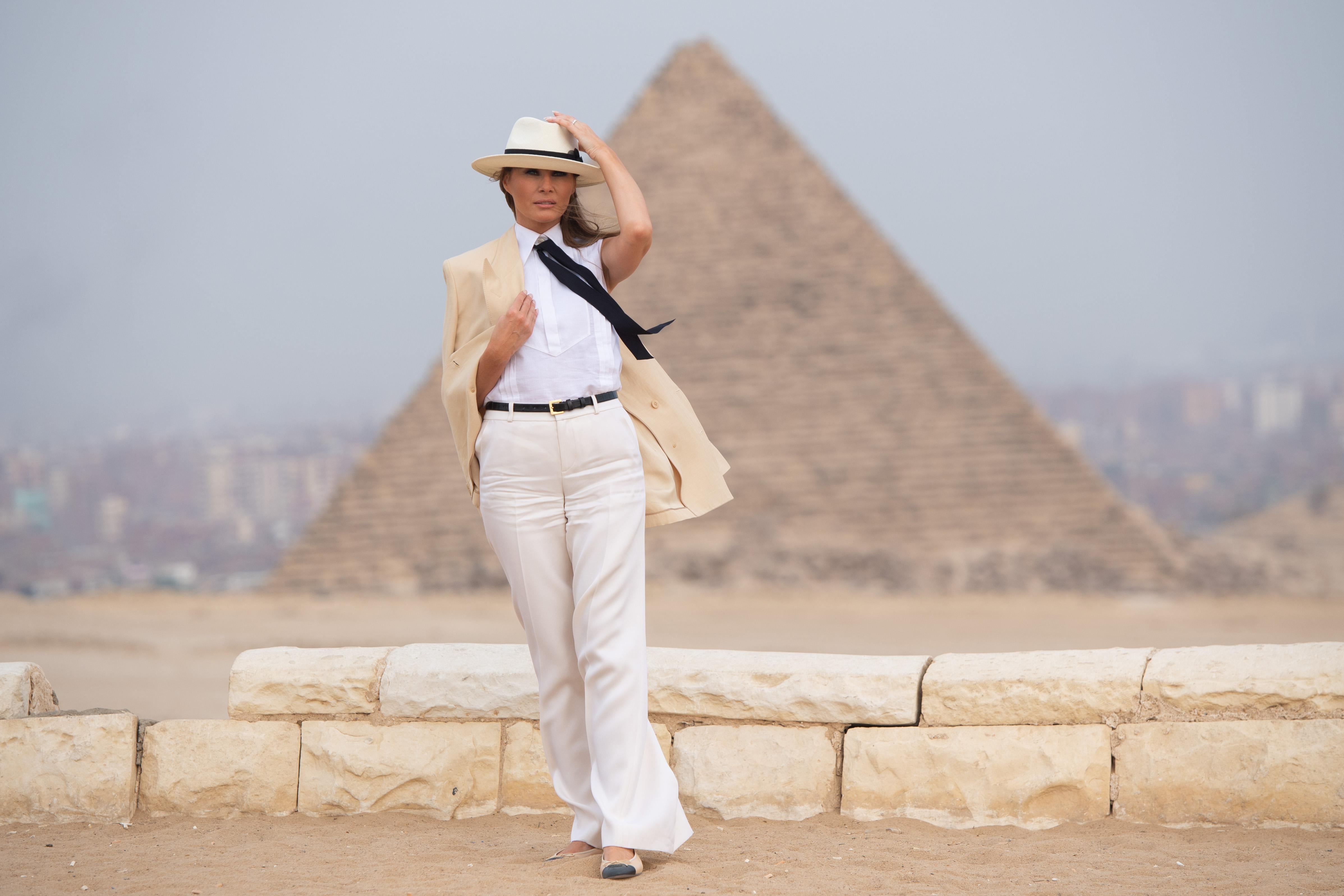 Melania Trump clasps her beige blazer with one hand and her cream fedora with the other in front of a gigantic pyramid. She wears a white shirt and long black tie.