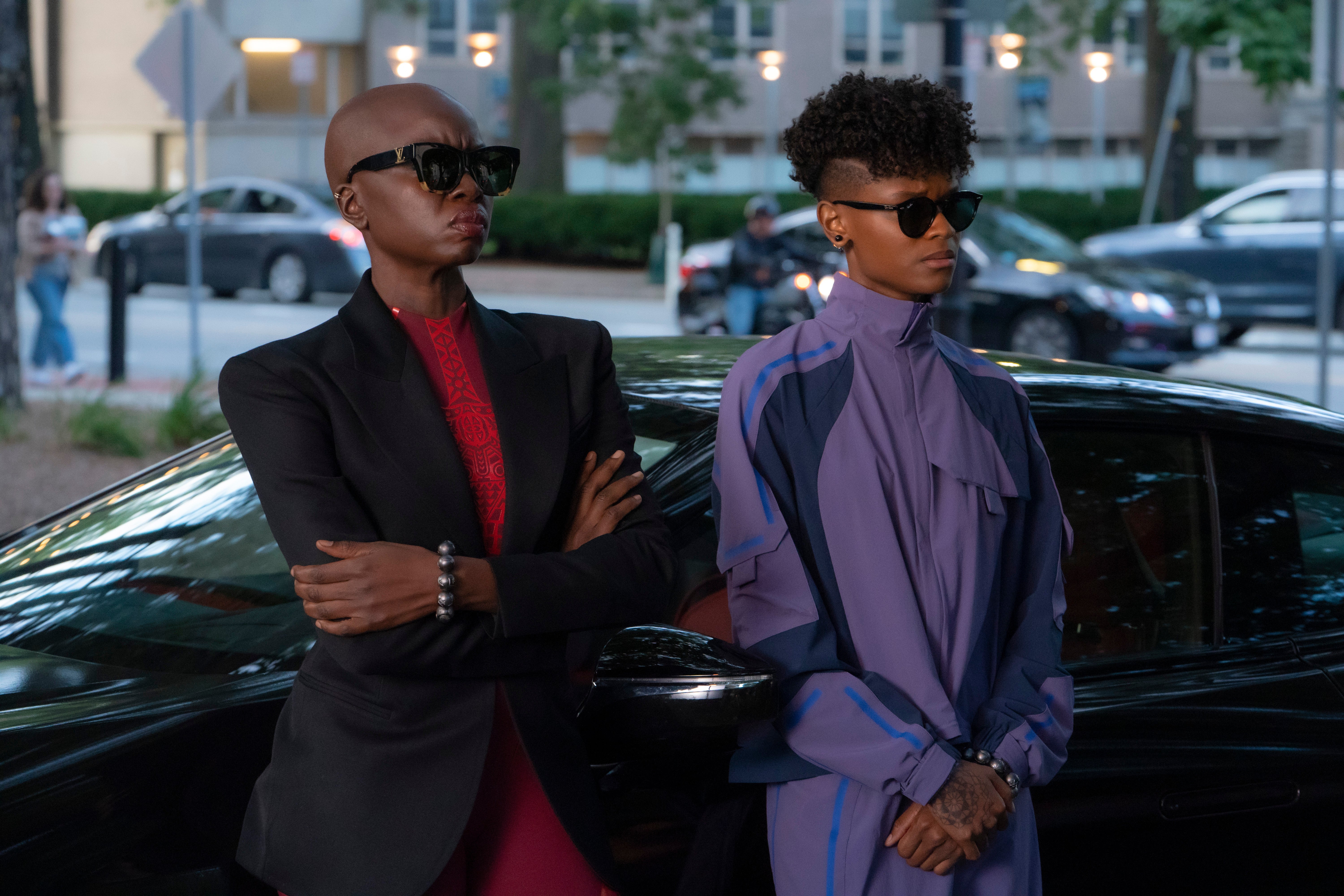Both lean back against a black sedan, looking stylish in black sunglasses. Okoye wears a black blazer over more traditional red outfit befitting of one of Wakanda's dora milaje, while Shuri wears a futuristic purple tracksuit.