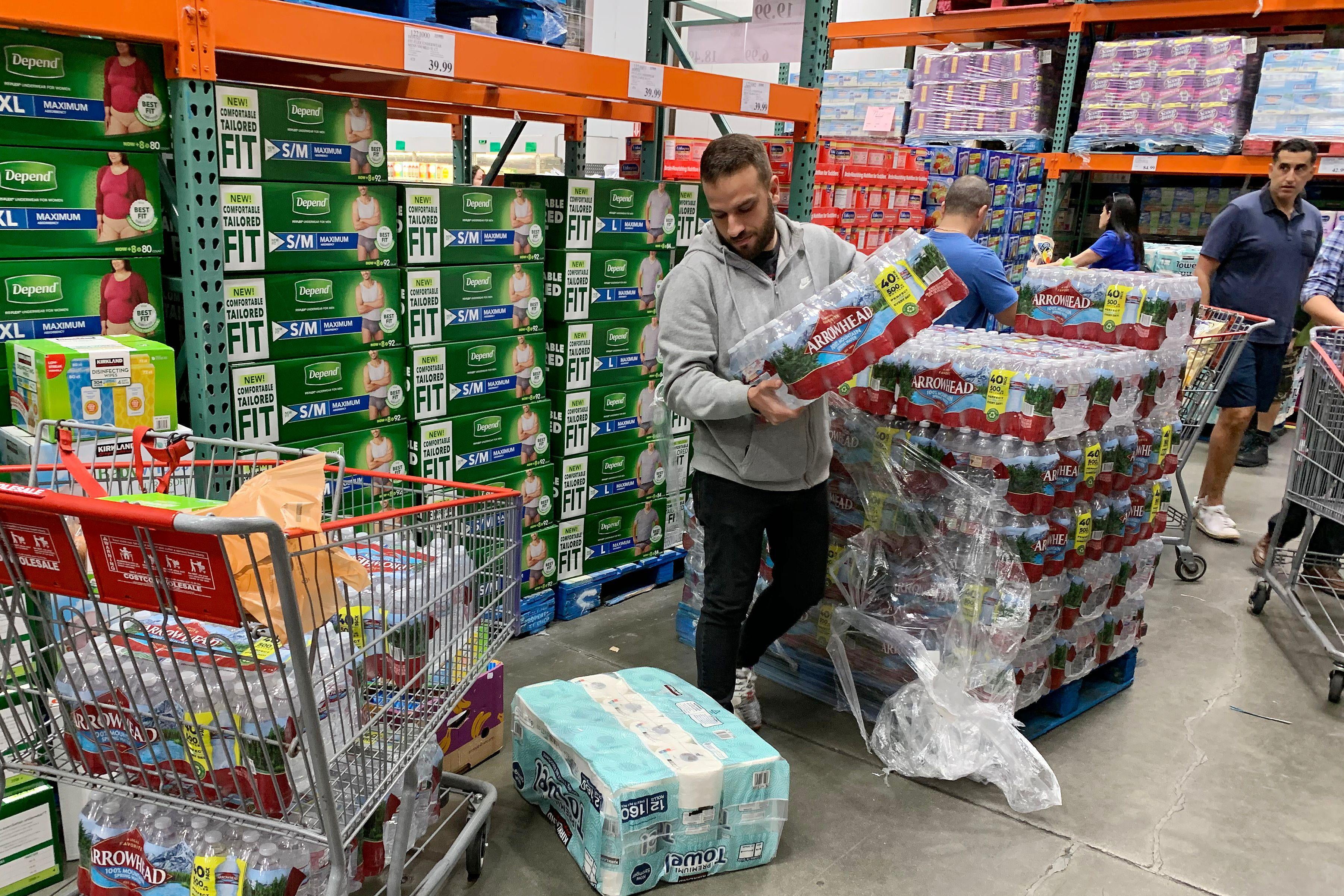 A man in a grocery store takes a case of water bottles from the top of a stack and looks at his shopping cart, which also has a case of water.