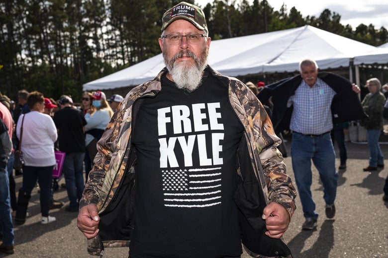 A man with a gray beard, eyeglasses, and a "Trump 2020" hat opens his camouflage jacket, revealing a shirt that says "Free Kyle" with a black-and-white American flag. People mill around behind him.