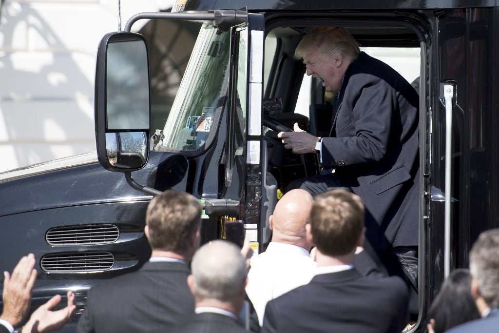 Trump sits in the cab of a semitruck and screams in front of a crowd of onlookers wearing suits.
