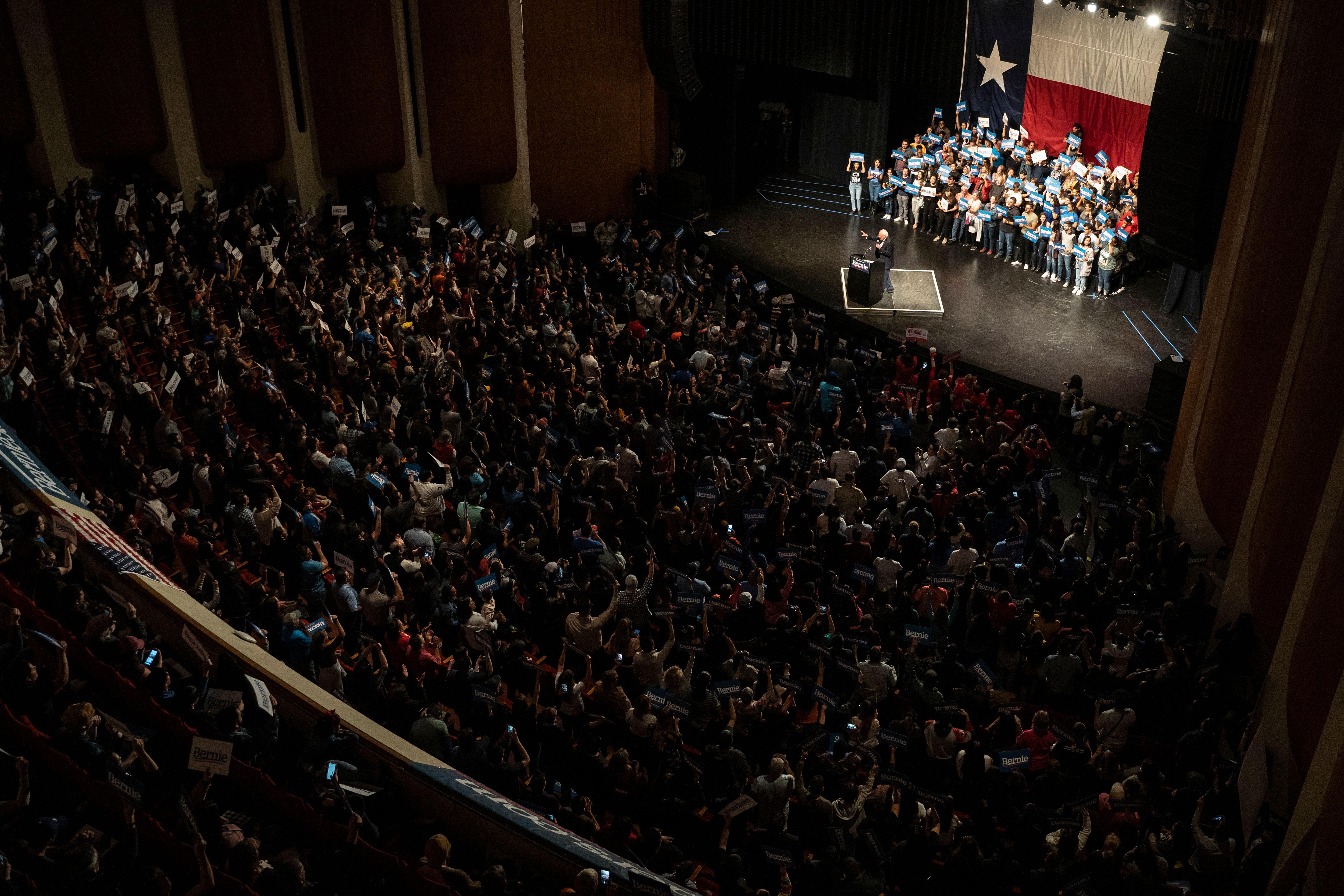 Seen from high up inside a theater, the tiny figure of Bernie Sanders stands on a brightly lit stage with a Texas flag at the back, addressing a full audience. 