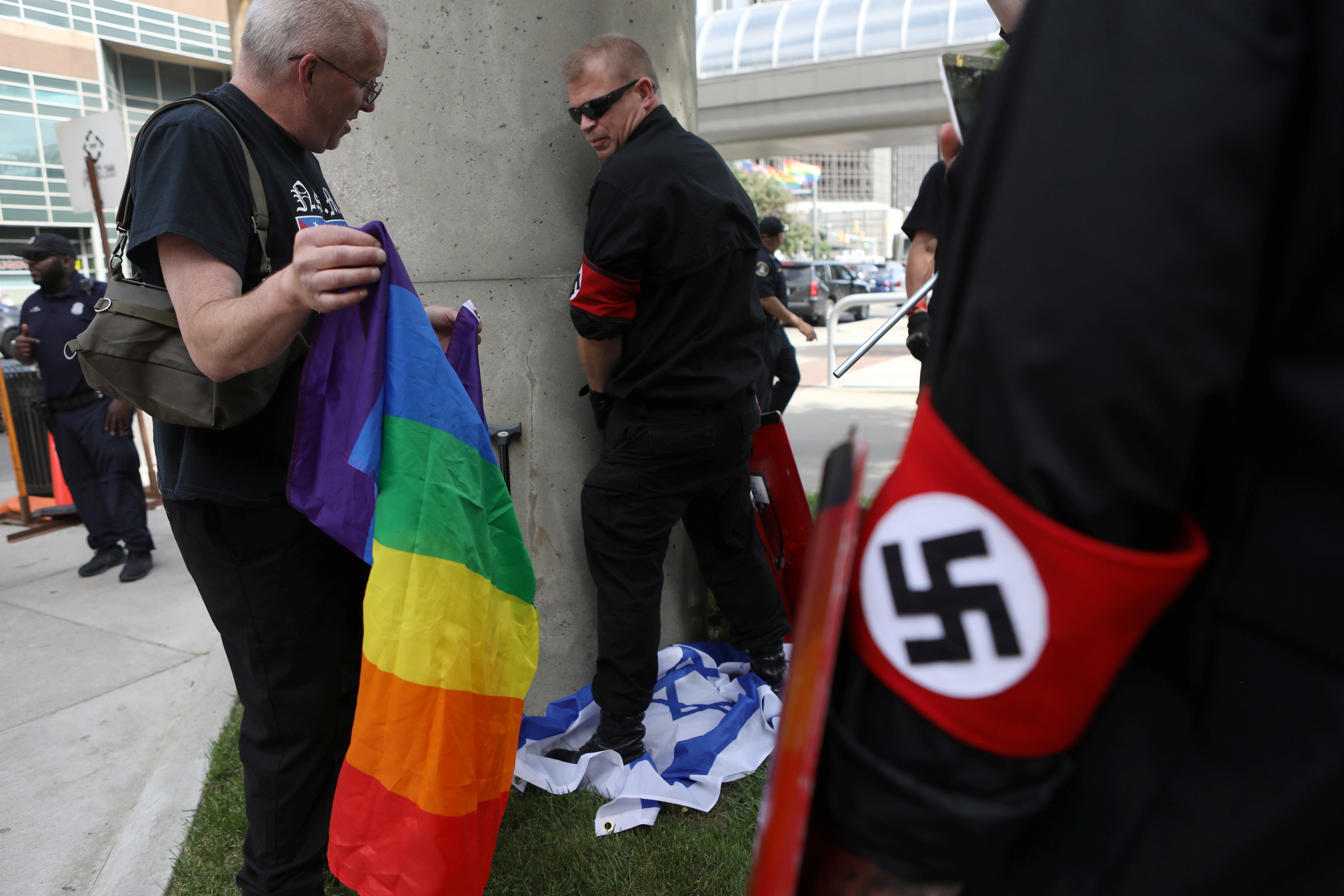 A group of neo-Nazis, one holding a rainbow pride flag, one stepping on an Israeli flag. A law enforcement official stands in the background.