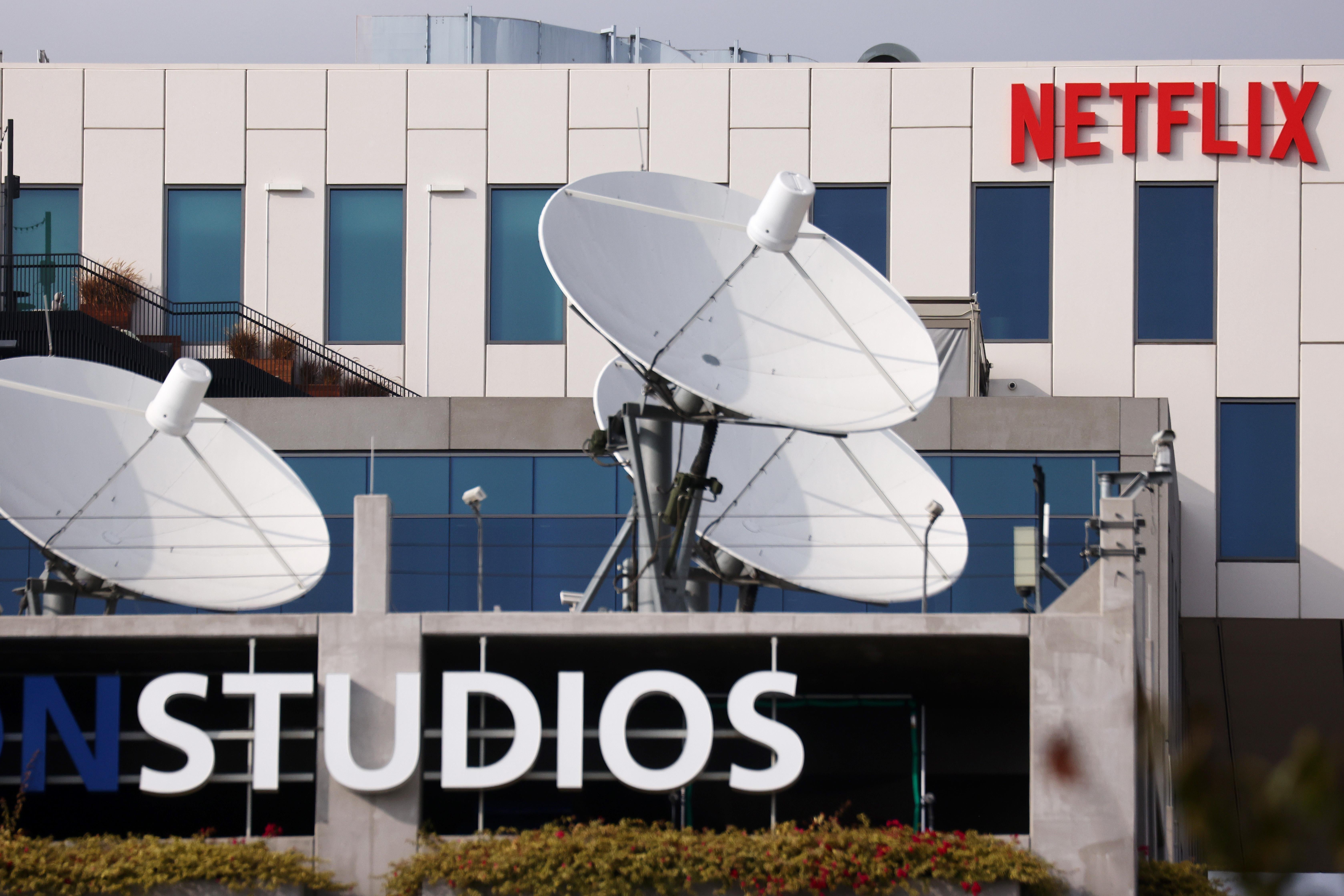 Exterior shot of a building with a Netflix sign in the background and a building with the word Studios on a logo and multiple satellite dishes on the roof in the foreground.