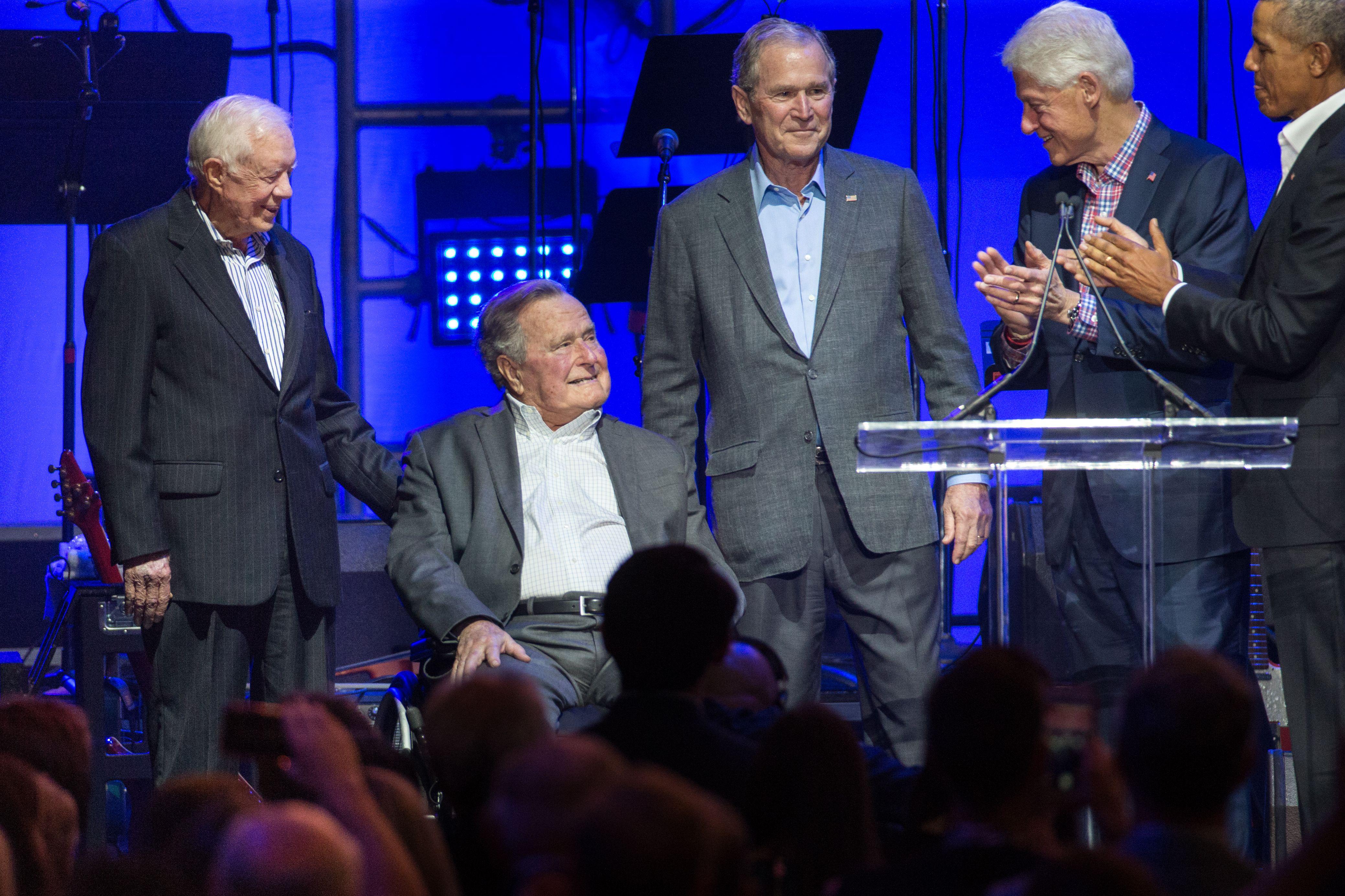 Jimmy Carter looks down toward George H. W. Bush and smiles on his left. To his right, his son George stands and smiles. To his left, Obama and Clinton applaud. They are all standing on a stage set up for a concert.