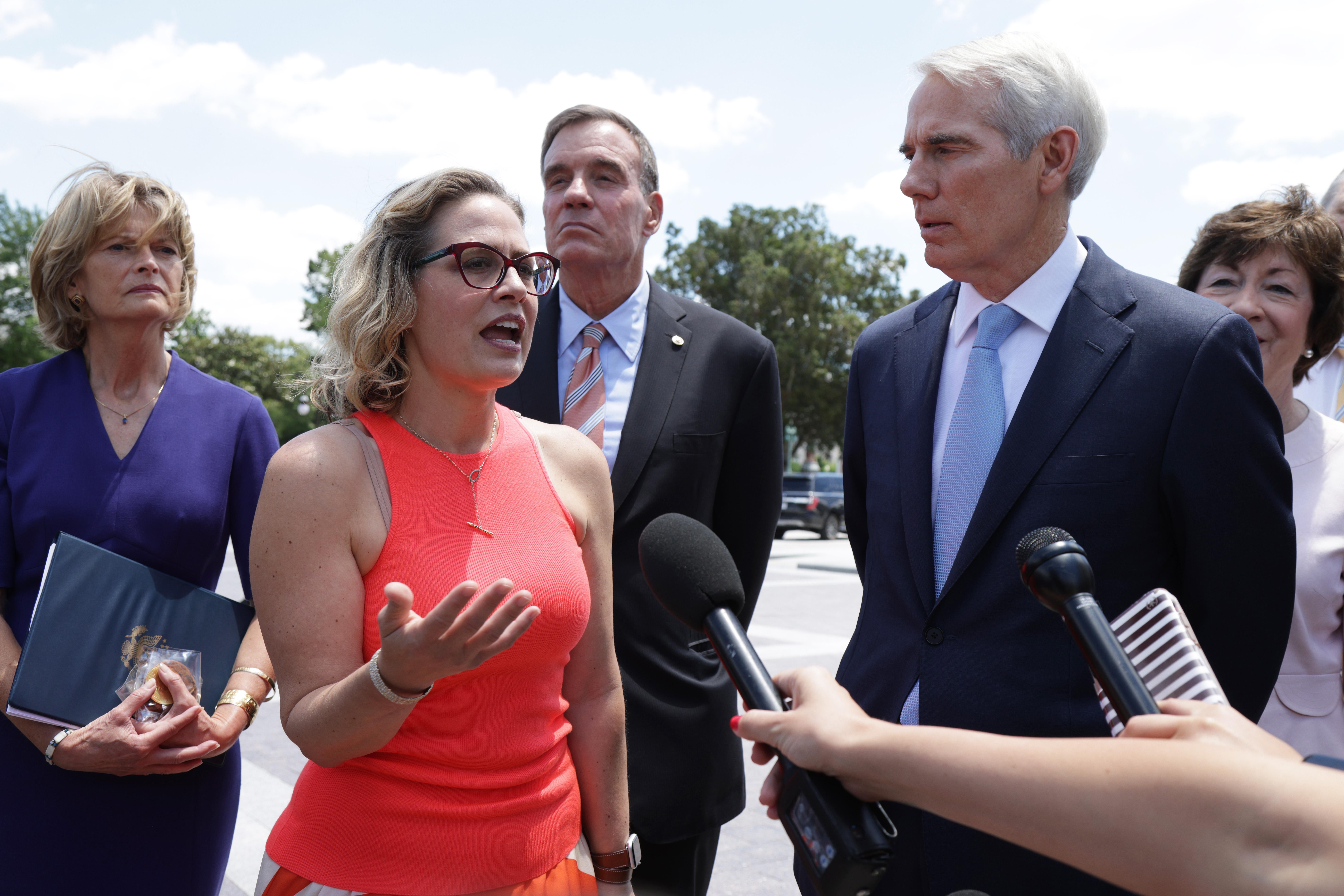 Sinema gestures with one hand as she speaks before a microphone held out by a reporter. Murkowski, Warner, Portman, and Collins stand around her outside.