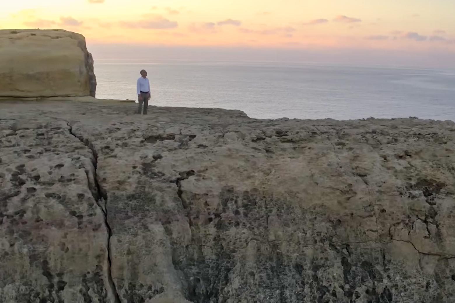 Journalist Graham Hancock, wearing a white shirt, stands on top of a rock formation at sunrise.