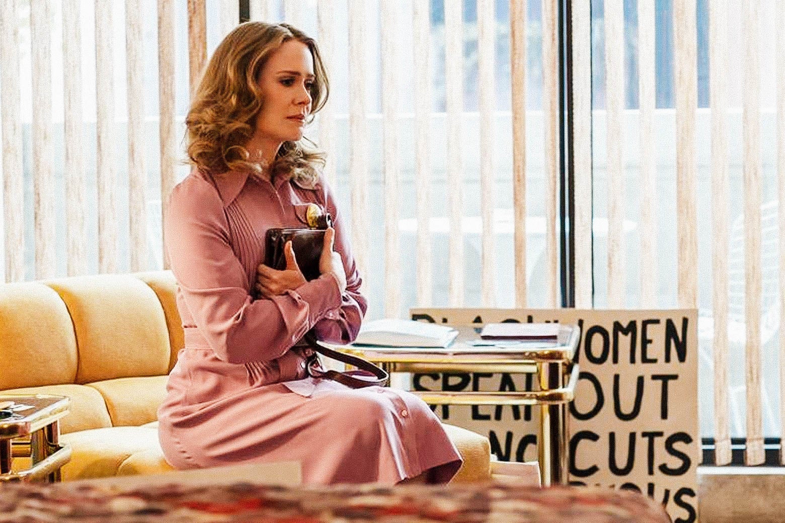 Sarah Paulson as Alice sitting on a sofa, clutching a book to her chest, looking conflicted.