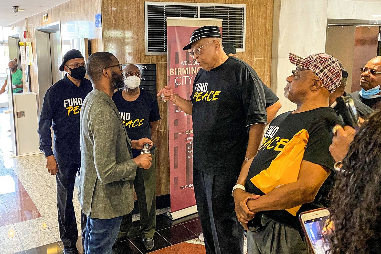 Men in Fund Peace T-shirts stand around a man in a blazer and jeans.