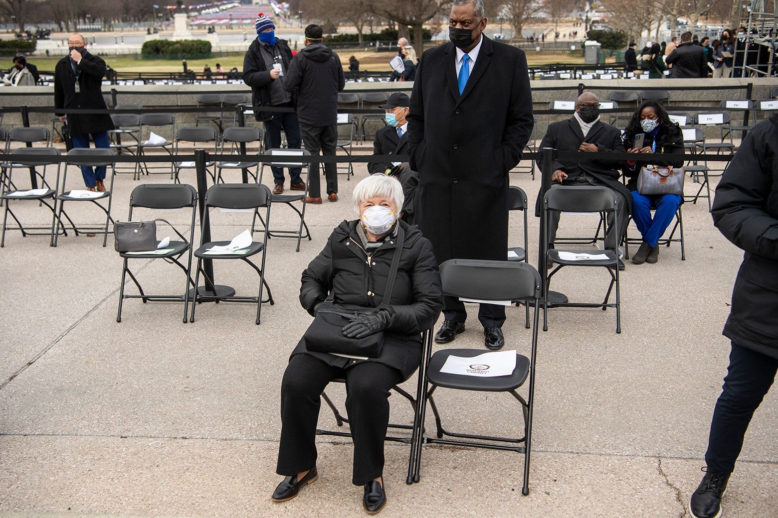 Janet Yellen sits in a folding chair in an all-black outfit with a white mask.
