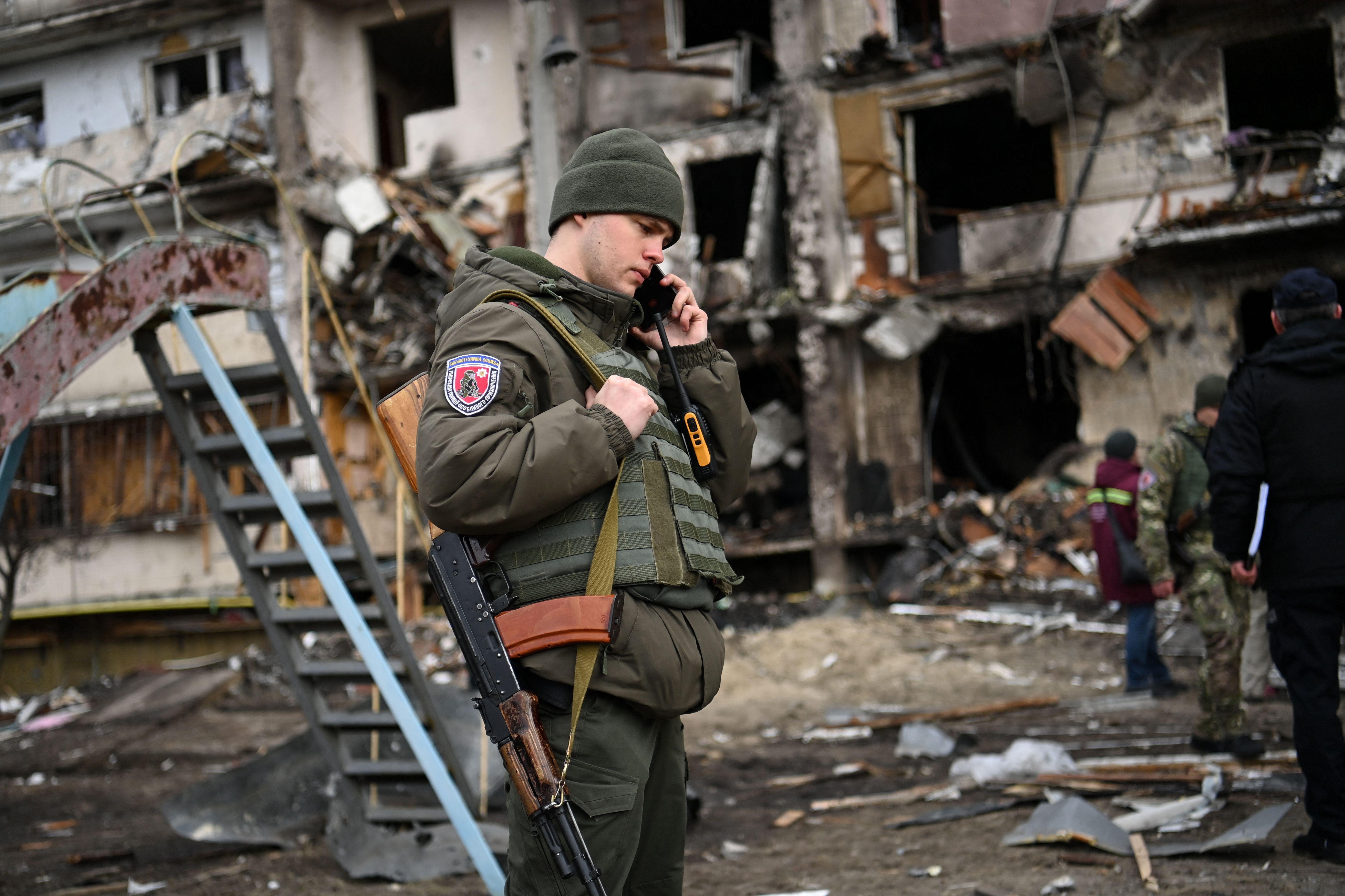 A man in uniform on the phone, his hand resting on the strap of his gun, in front of ruined buildings.