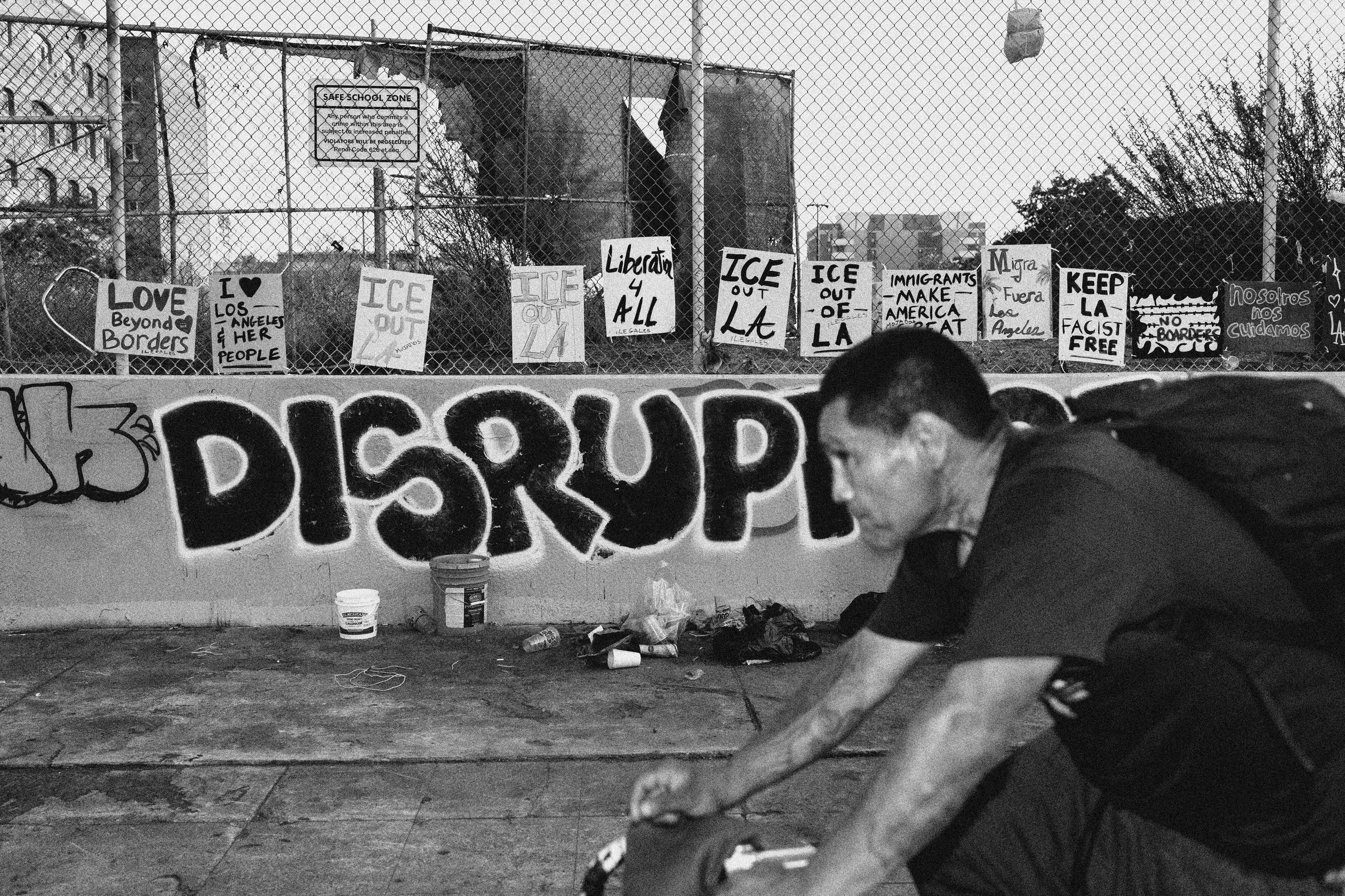 A man rides a bike past a wall with graffiti that says DISRUPT and that is lined with anti-ICE protest signs like "ICE out of LA" and "Love Beyond Borders"