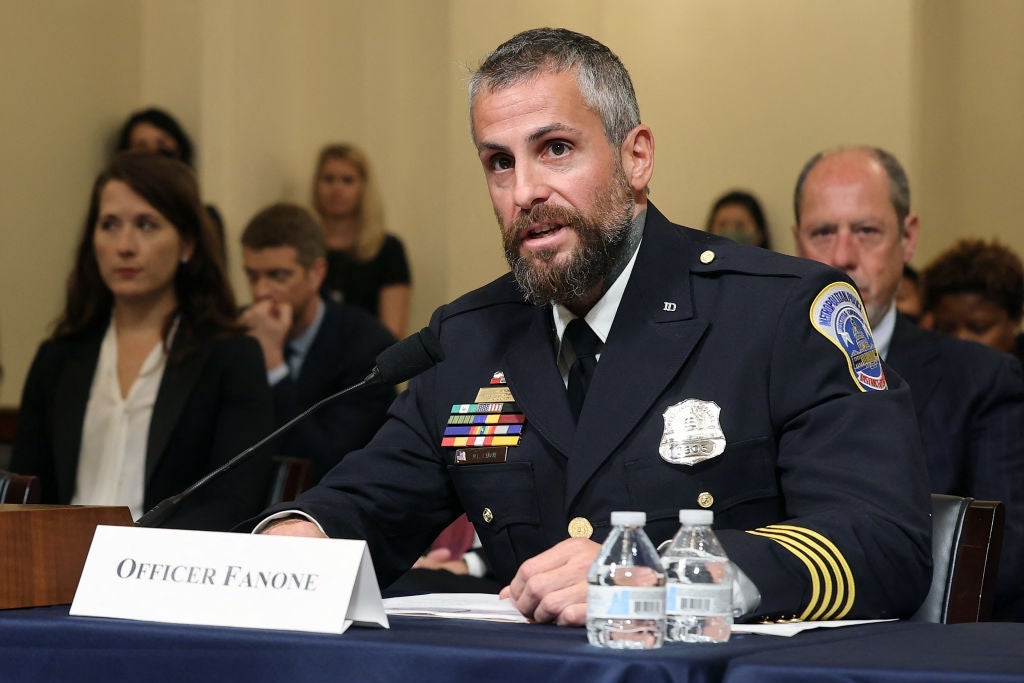A bearded police officer in dress uniform speaks into a microphone while seated behind a desk in a hearing room.