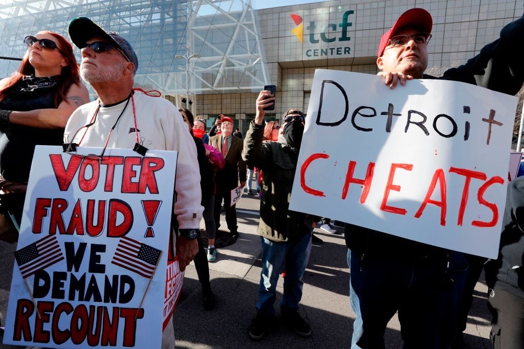 Older white men hold signs reading "DETROIT CHEATS" and "VOTER FRAUD! WE DEMAND RECOUNT."