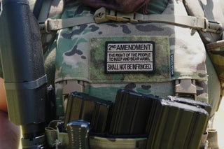 A man's camo vest, stuffed with weapons and ammunition, reads the text of the Second Amendment.