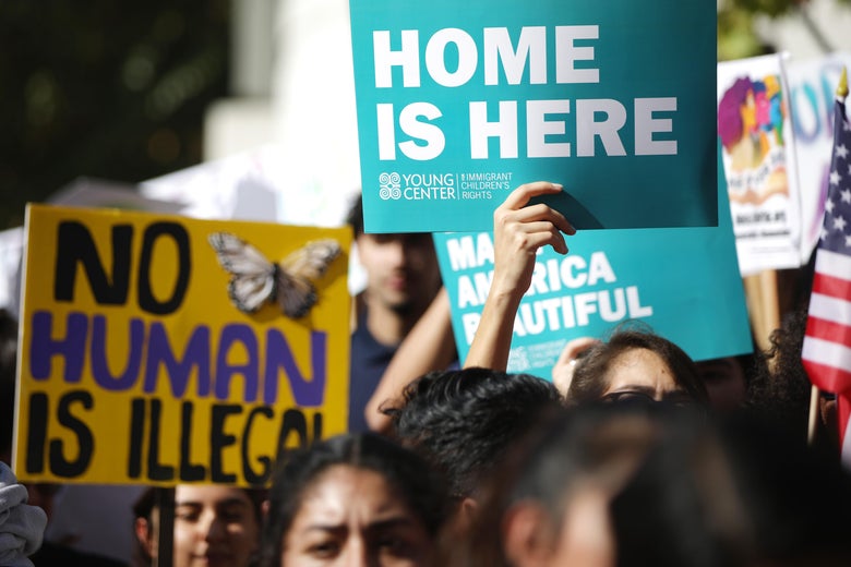 A crowd of people hold up signs saying No human is illegal and home is here.
