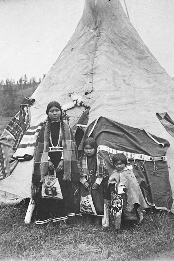 A young woman and two children stand in front of a skin tepee.