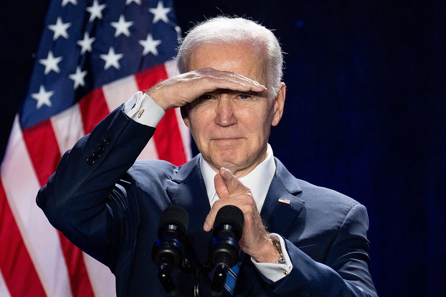 Joe Biden at a podium pointing and covering his eyes from a glare.