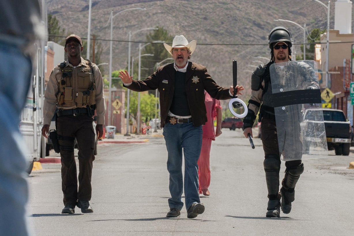 A sheriff walks down the center of a New Mexico street toward the camera, deputies on each side. His hands are raised, as if to try to calm the situation down. On the left edge of the frame, we can see the leg of a man's jeans, facing him down.