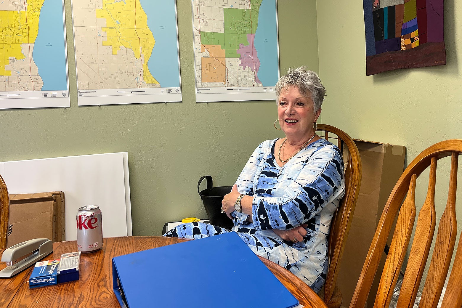 A woman in a blue and white top sitting at a table with her arms folded in a light-green room with maps on the wall.