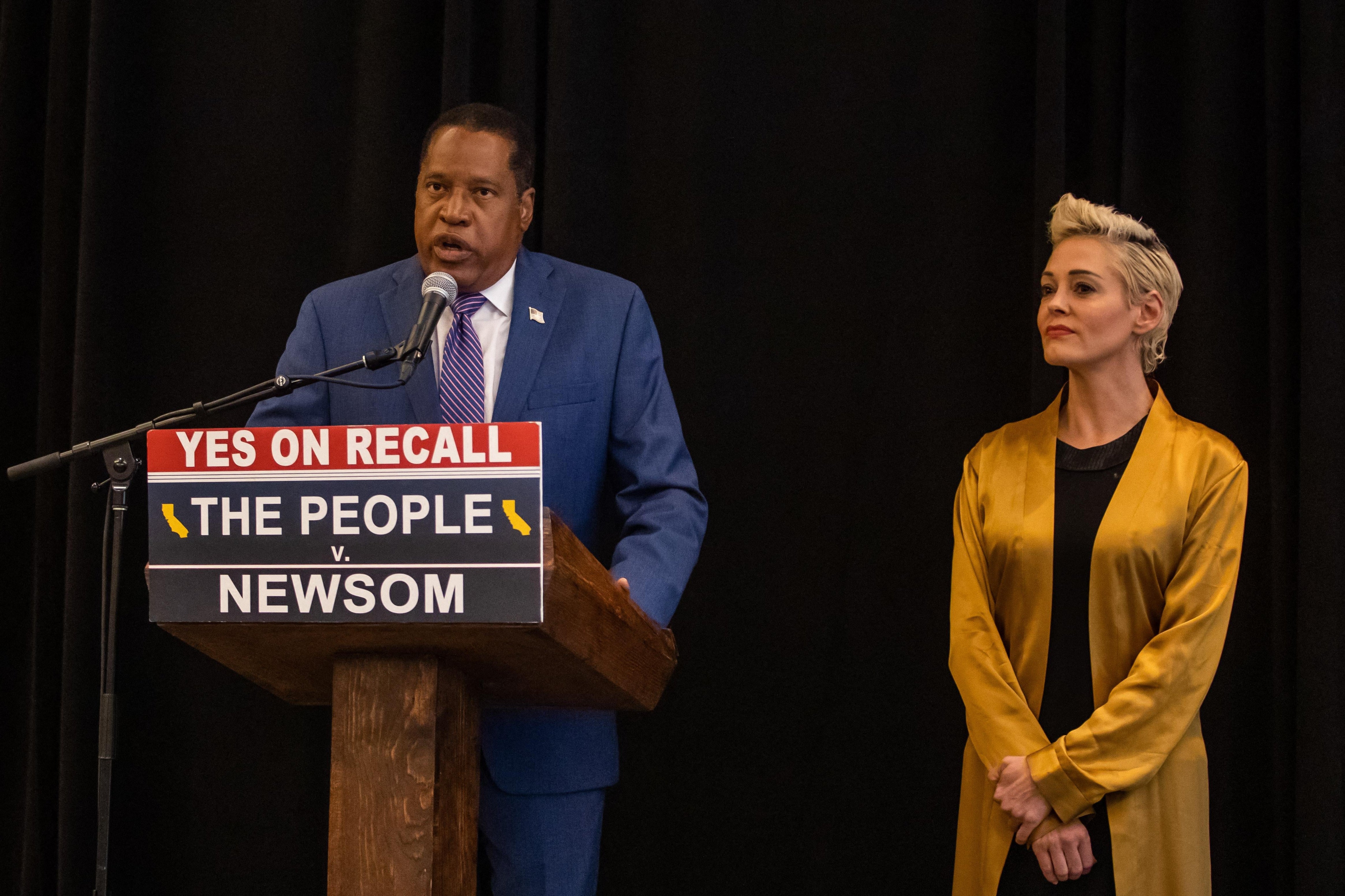 Larry Elder speaks into a microphone at a podium with a sign that says "Yes on Recall. The People v. Newsom." Rose McGowan stands beside him.