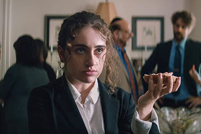 A young woman in a blazer and white button-down holds an appetizer in one hand.