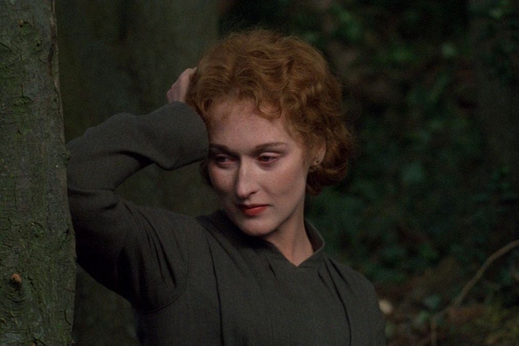 A young Meryl Streep leans against a tree in the woods while staring off into the distance. 