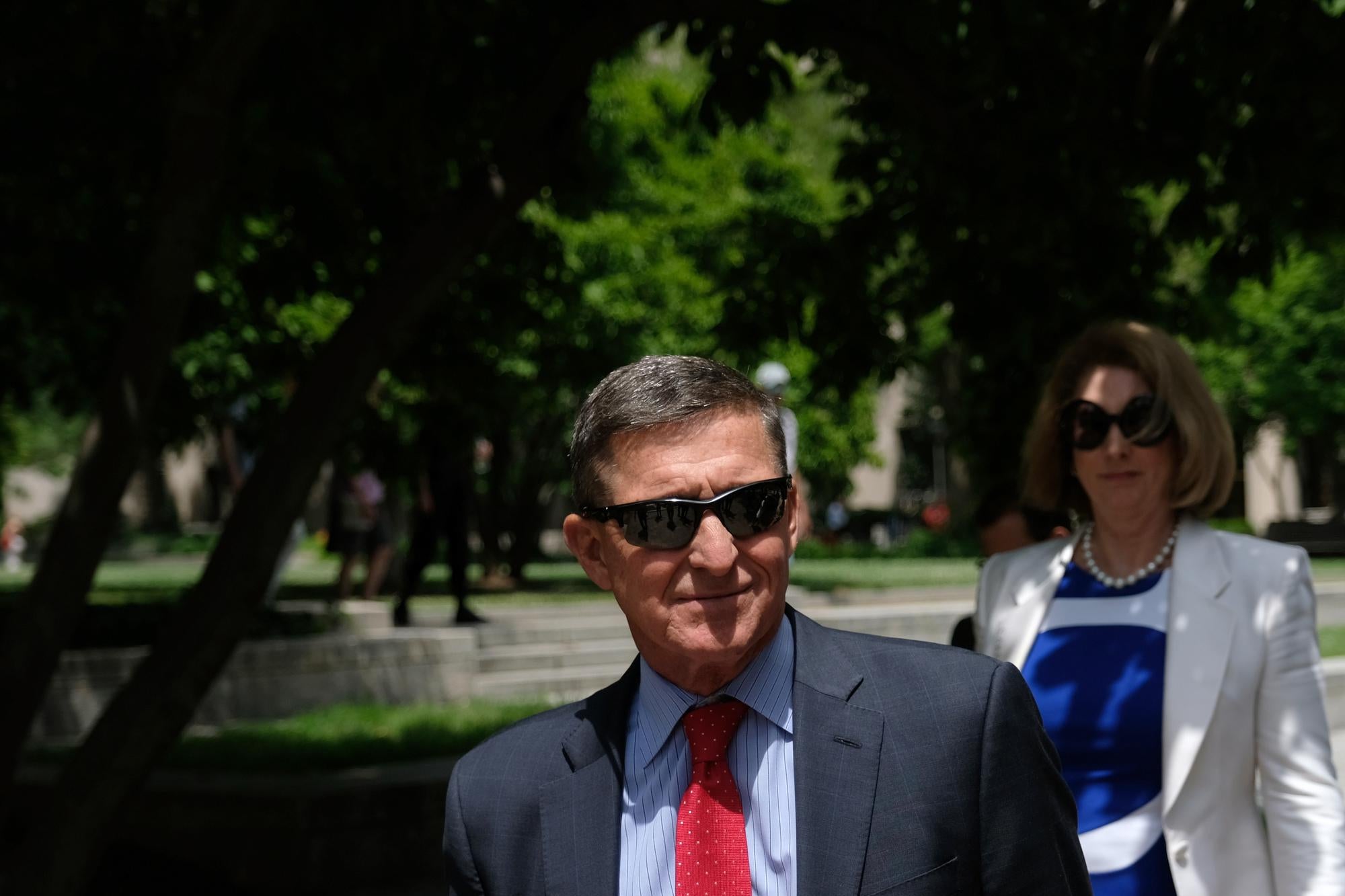 Michael Flynn wearing sunglasses and a suit and tie, with a woman in sunglasses walking behind him.
