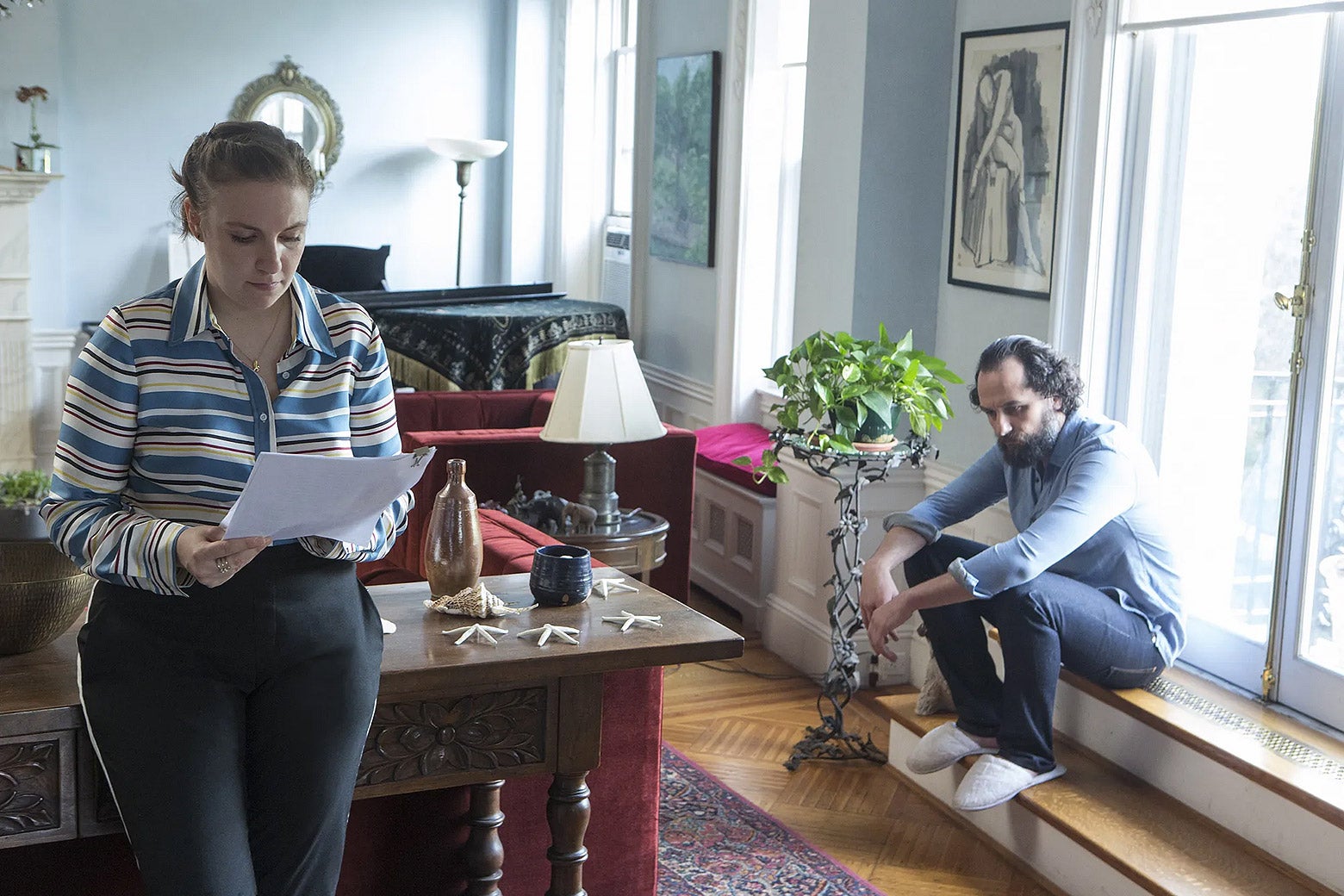 A white twentysomething woman wearing a blue striped shirt stands in a fancy apartment reading a piece of paper. Behind her, a middle-aged white man in a blue button-down and jeans sits by a window and stares at the floor.