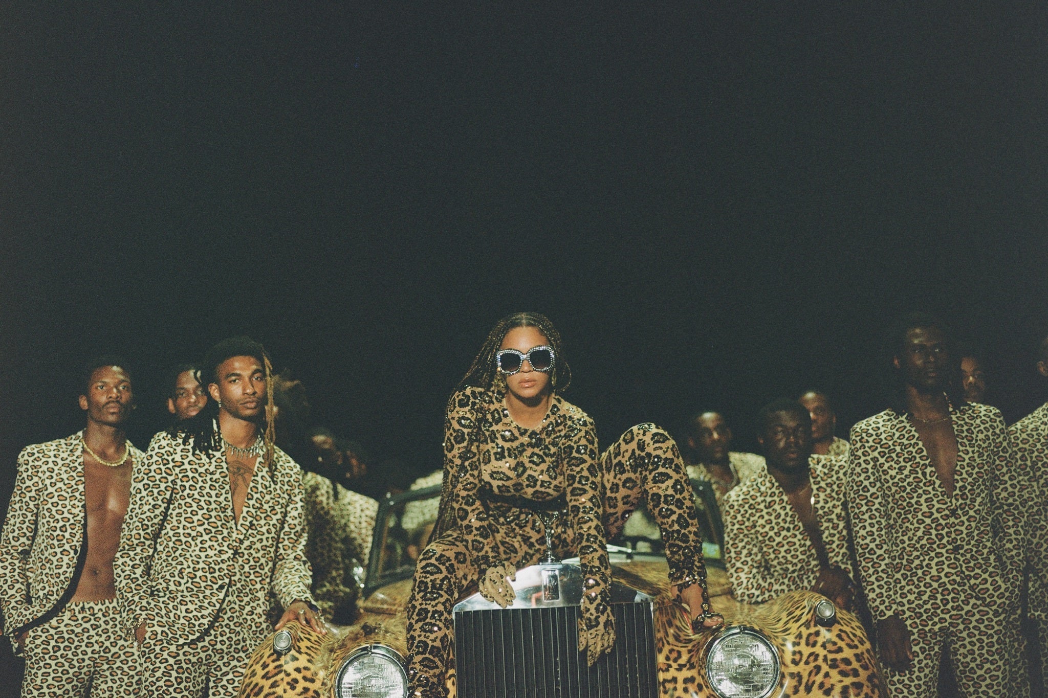 Beyoncé sits atop the grill of a vintage-looking, leopard-print automobile, surrounded by Black men wearing leopard-print blazers and leopard-print pants. She wears sunglasses, braids, and leopard print.