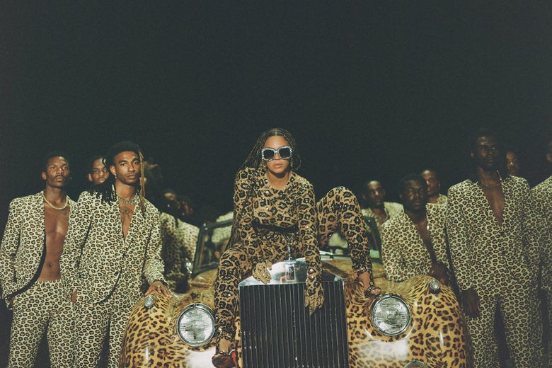 Beyoncé sits atop the grill of a vintage-looking, leopard-print automobile, surrounded by Black men wearing leopard-print blazers and leopard-print pants. She wears sunglasses, braids, and leopard print.