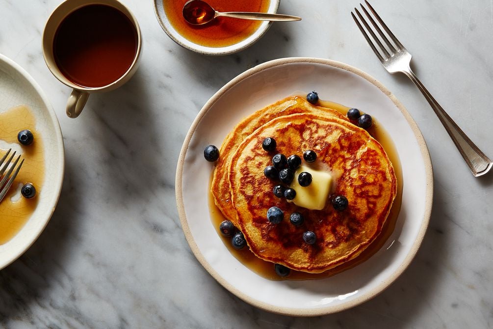 Stack of pancakes topped with blueberries, a pat of butter, and syrup, next to a fork, a cup of coffee, a small dish of syrup, and a finished plate of pancakes on a marble surface