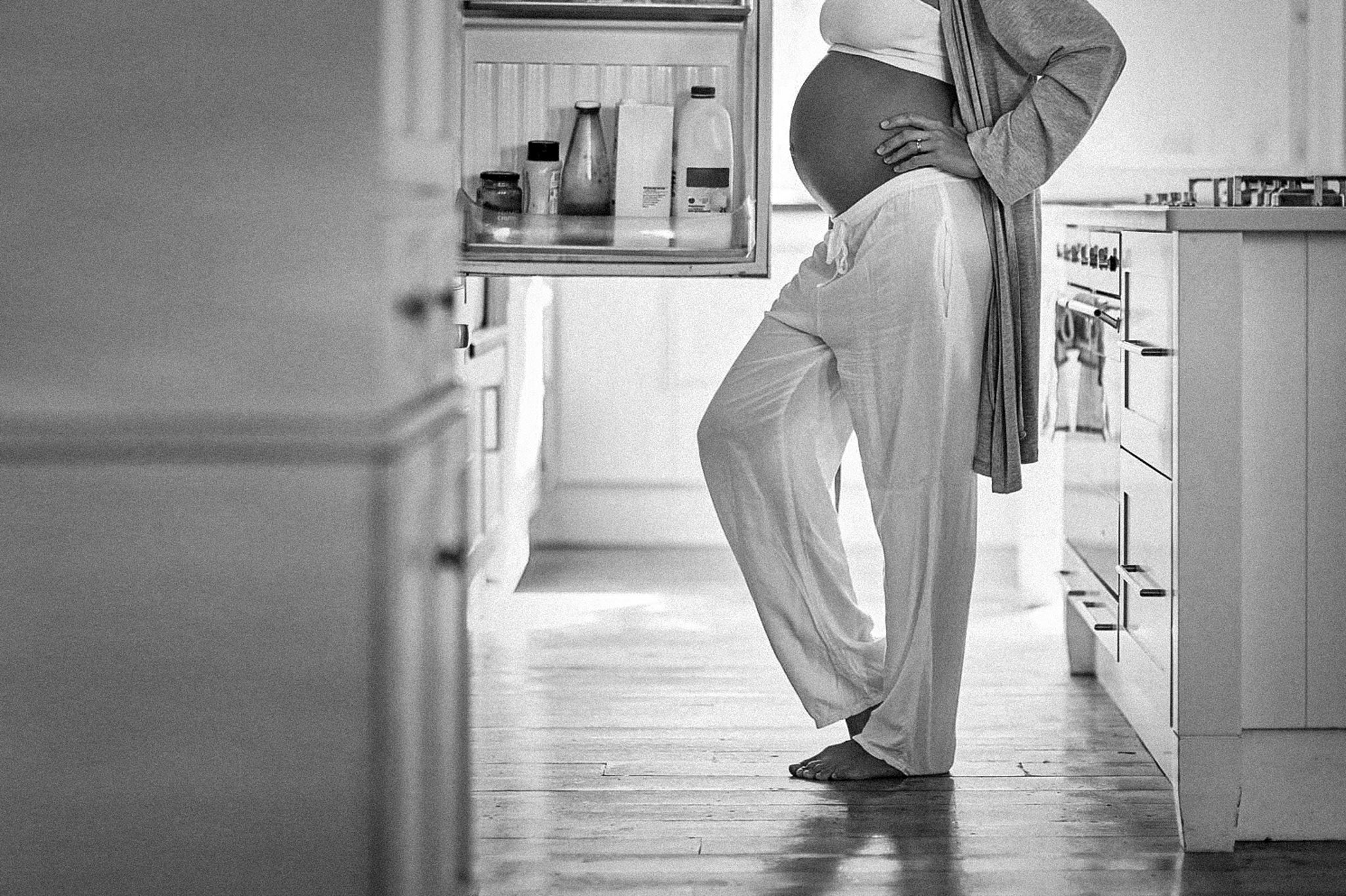 A black-and-white photo of a pregnant woman standing barefoot in front of a refrigerator.