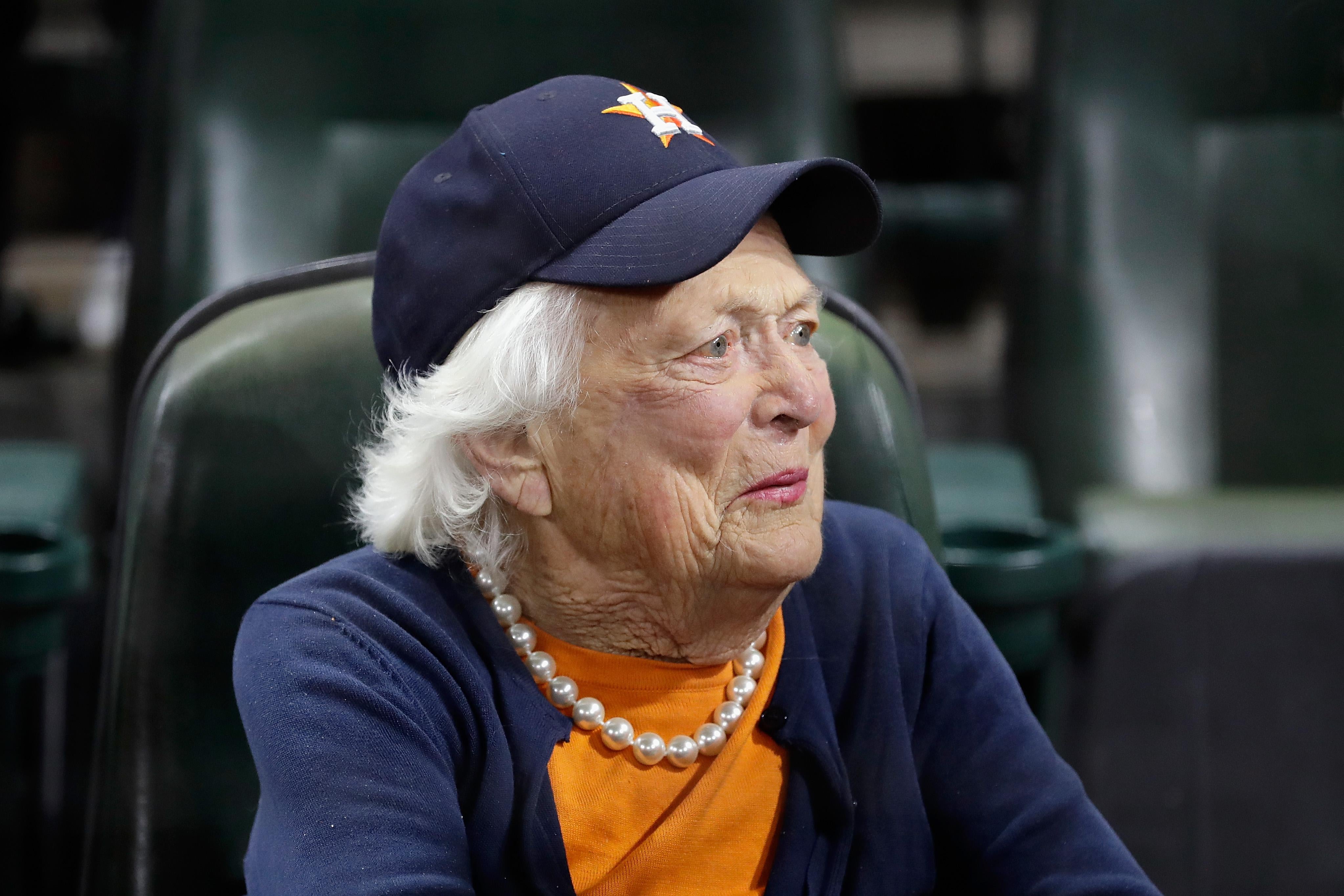 Barbara Bush, wearing a T-shirt, pearls, and a Houston Astros baseball cap, is seen sitting in stadium seating.